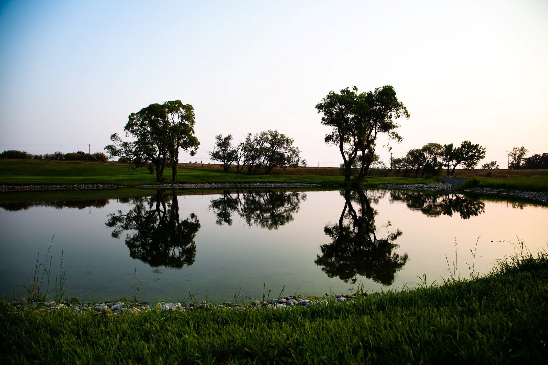 Pond with calm water reflecting trees and grassy banks at Hell's Creek Golf & Footgolf Course.