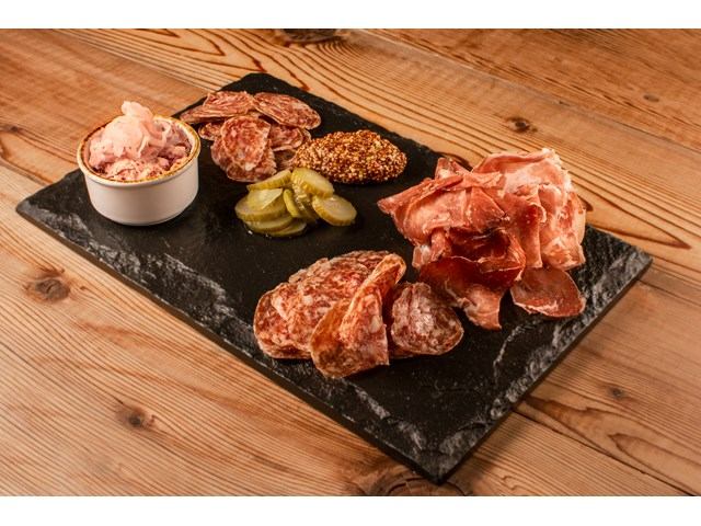Charcuterie board with sliced cured meats, pickles, mustard and spread on black slate.