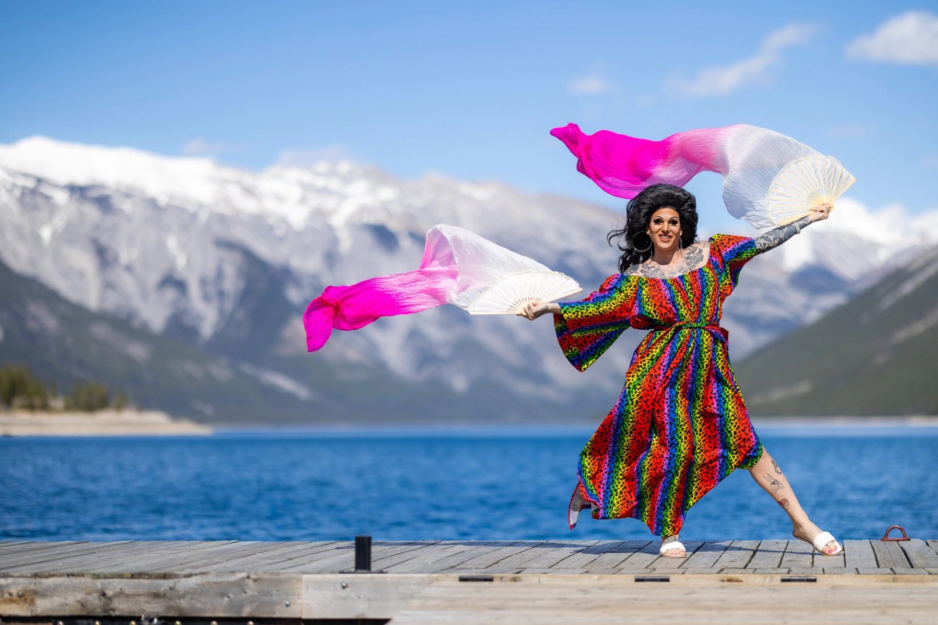 Person in rainbow dress dances with fans on dock, lake and snowy mountains behind.