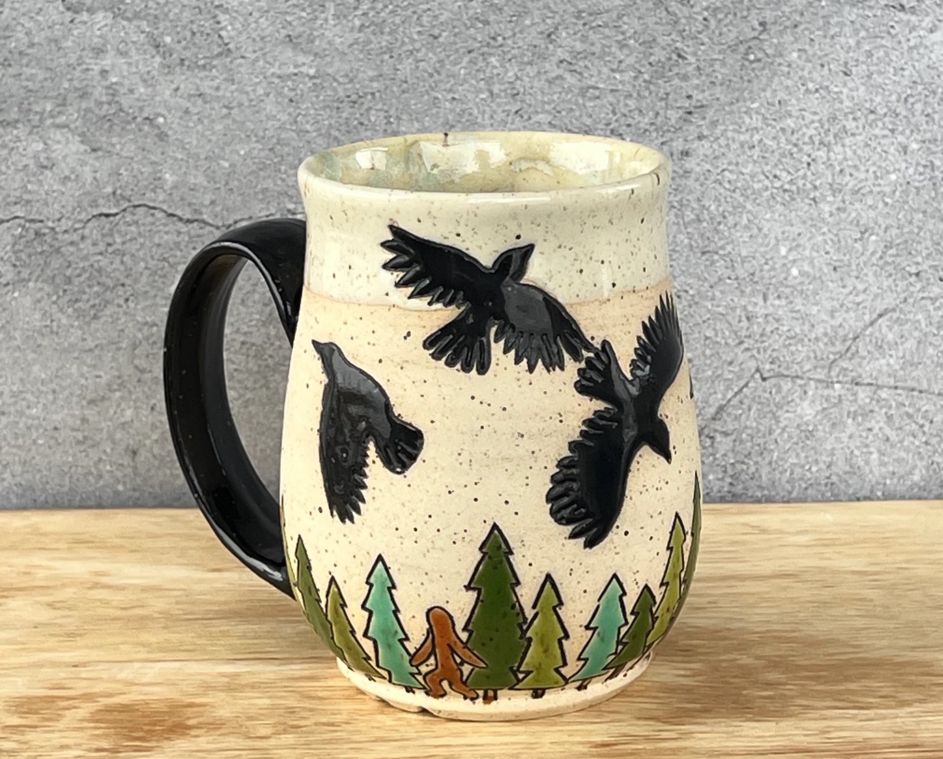 A handmade ceramic mug featuring a design with black birds and green trees at the base. 