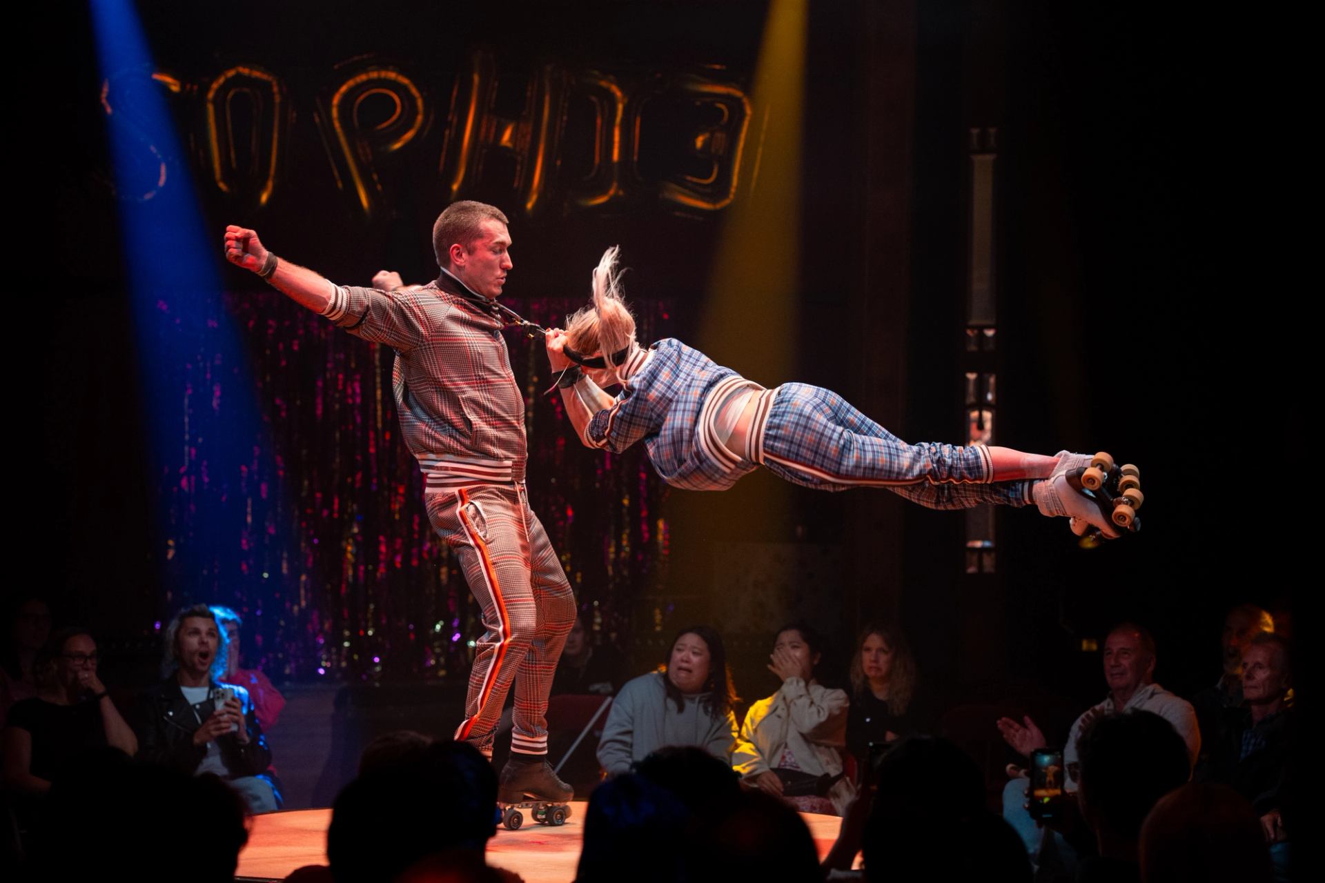 Roller skater spins partner horizontally by the neck on stage before seated audience.