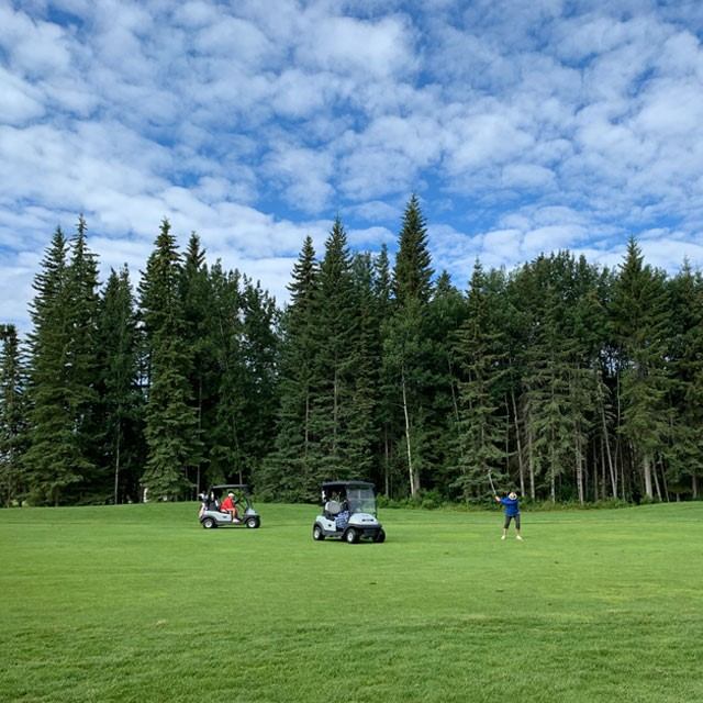 Golfers and carts on an open fairway at The Dunes Golf & Winter Club bordered by trees and blue sky.
