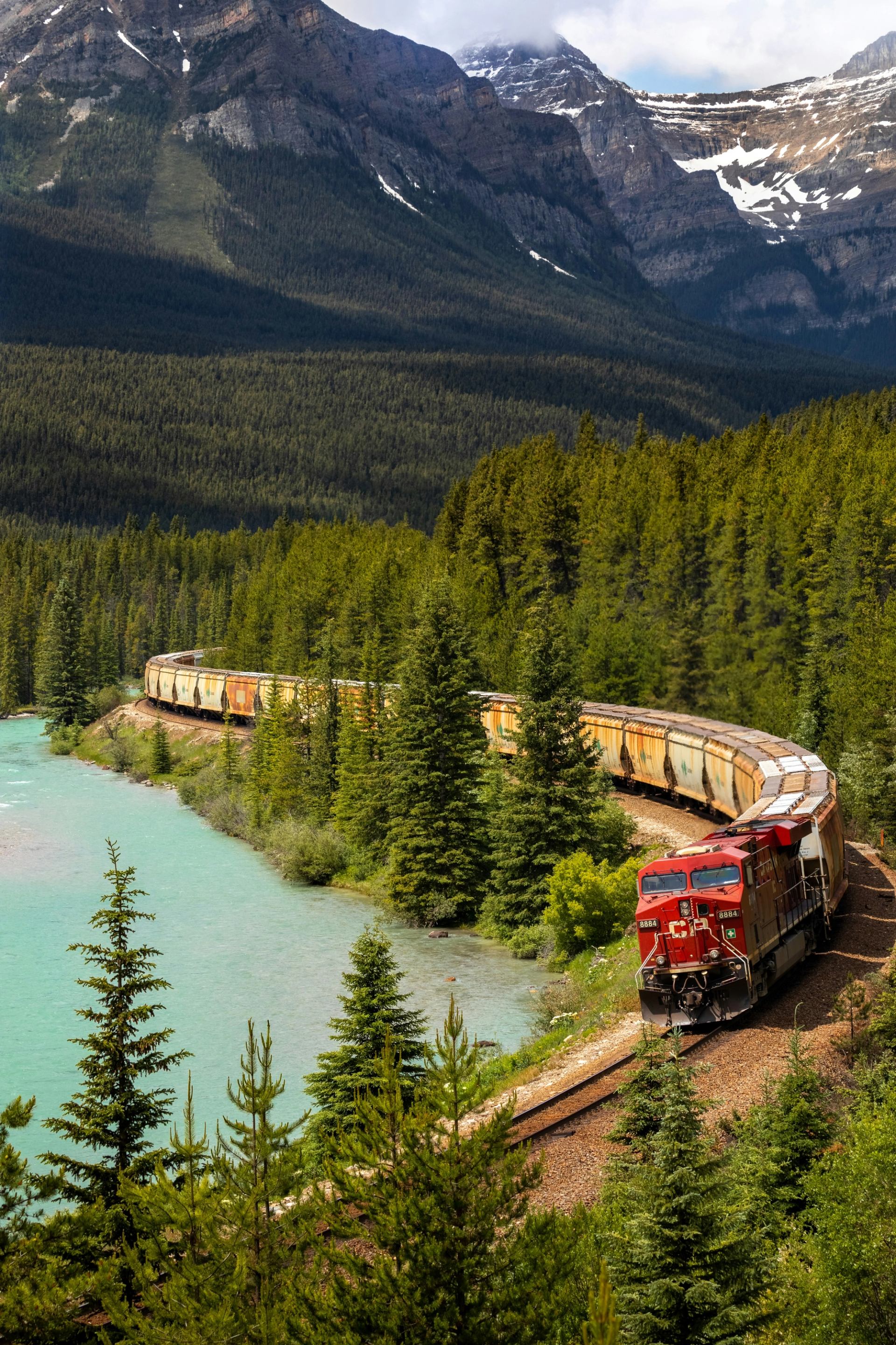 Freight train curves beside turquoise river through dense evergreen forest and steep mountains.