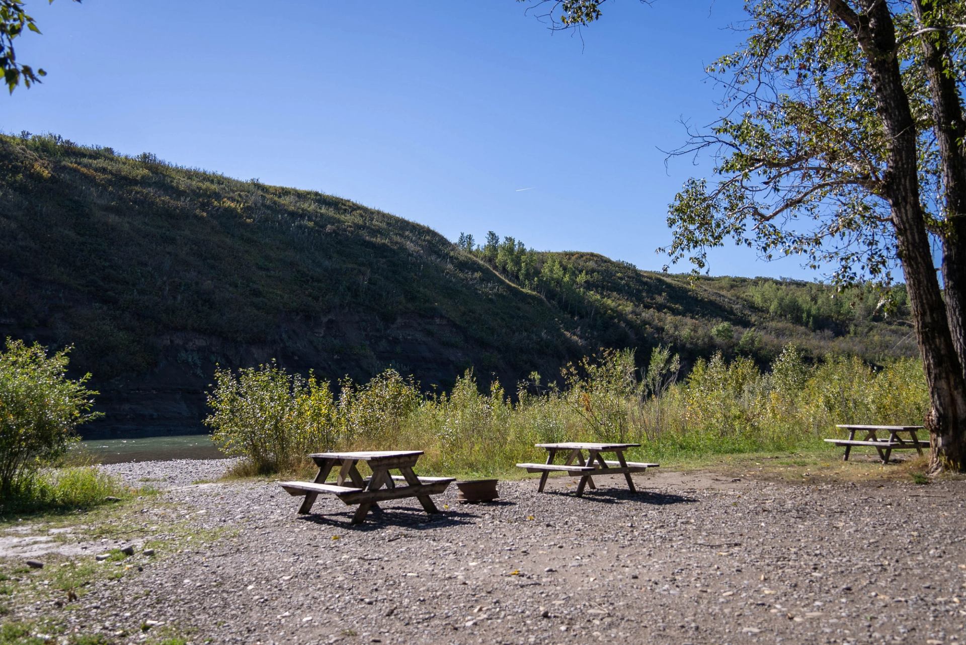Three picnic tables and a fire pit at a campsite with a steep, grassy hill in the background.