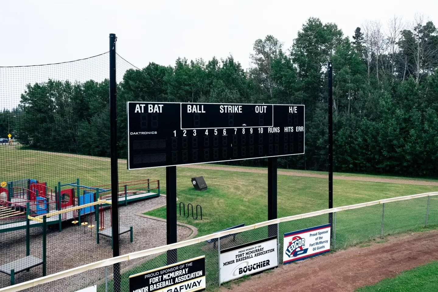 Baseball field scoreboard above chain-link fence, playground and trees in background.