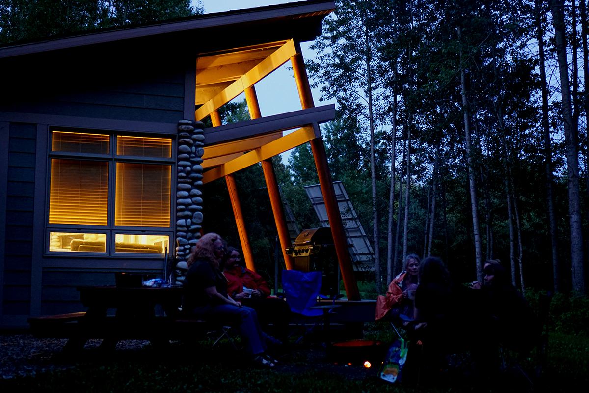 A group of women sit around a campfire, with The Nest Comfort Camping in the background.