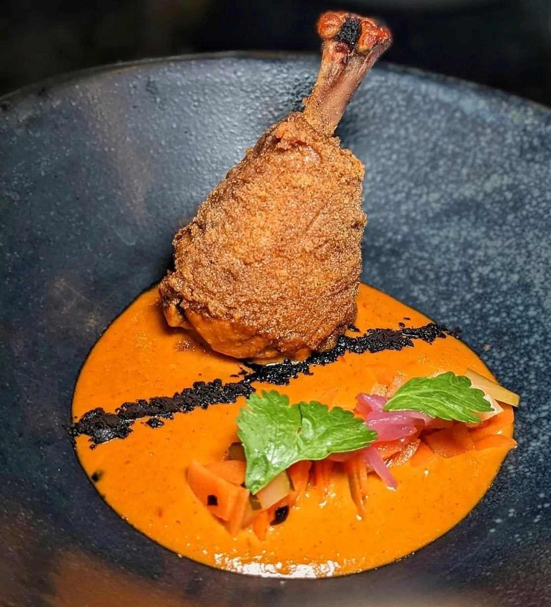 Fried chicken leg plated on orange sauce with garnish in a black bowl.