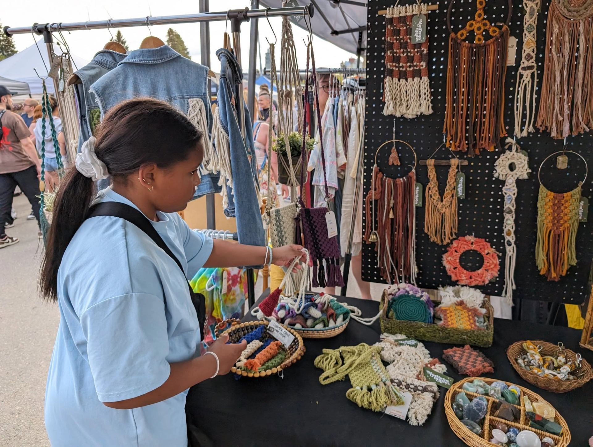Person browsing macrame wall hangings and handmade crafts at an outdoor market booth.