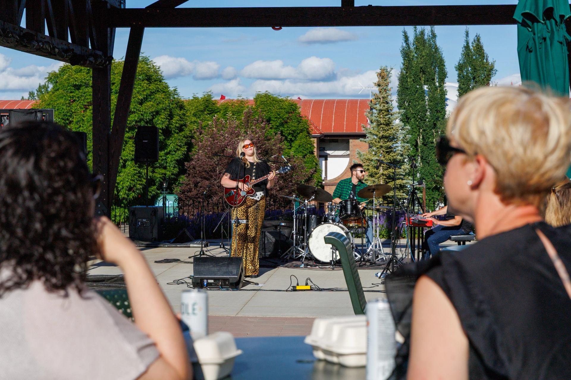 A musician performs on an outdoor stage as spectators watch from shaded seating.