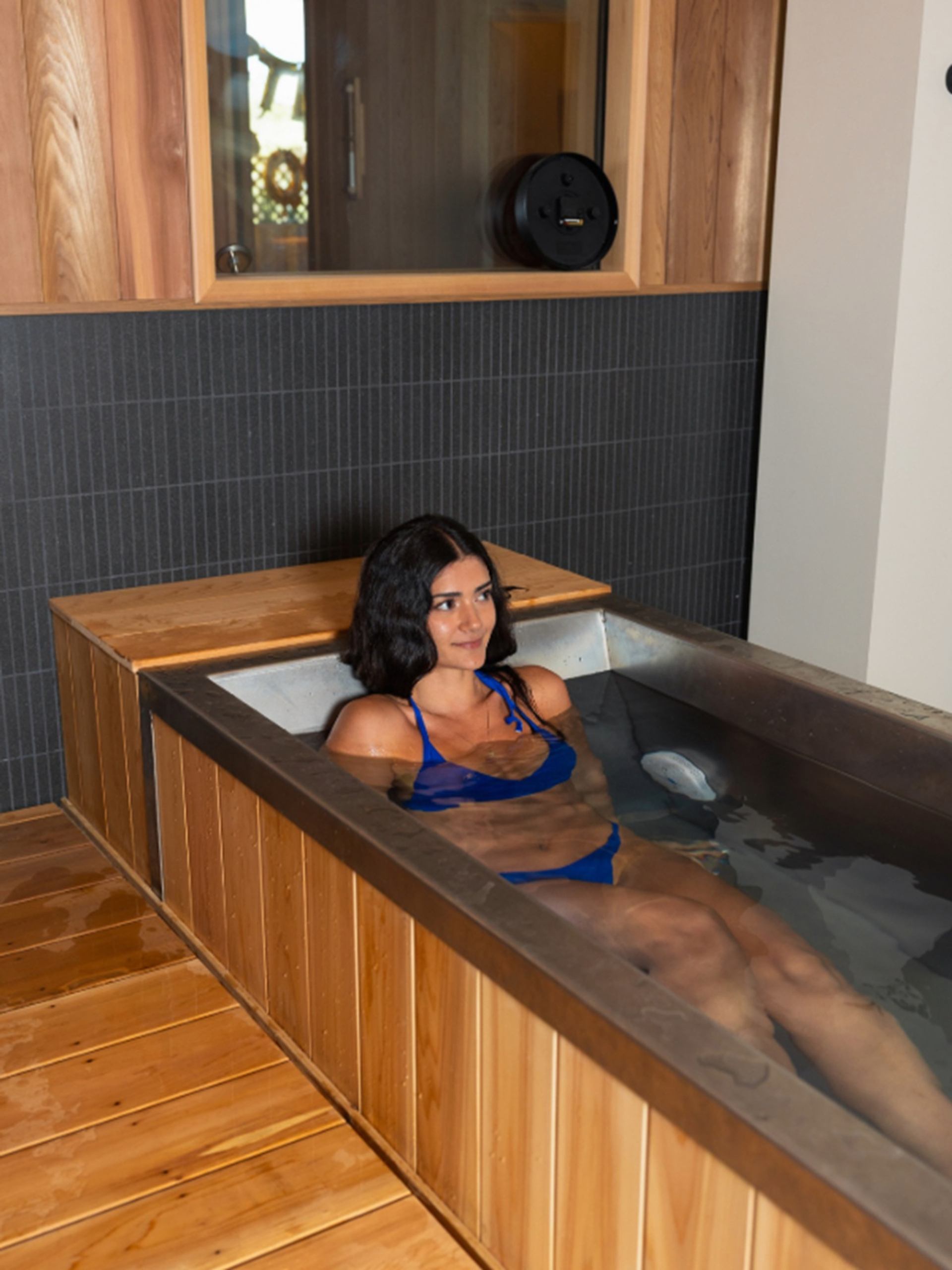 Person in blue swimsuit relaxing in a wooden soaking tub inside a modern spa room.