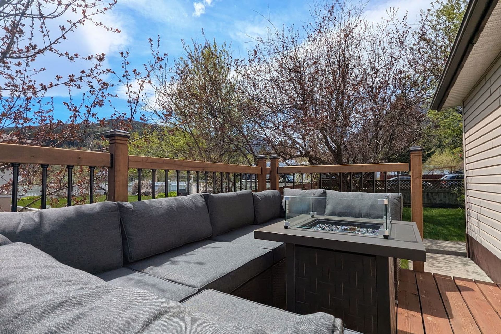 Outdoor wooden deck with a grey L-shaped sectional sofa, fire pit table, and view of distant hills.