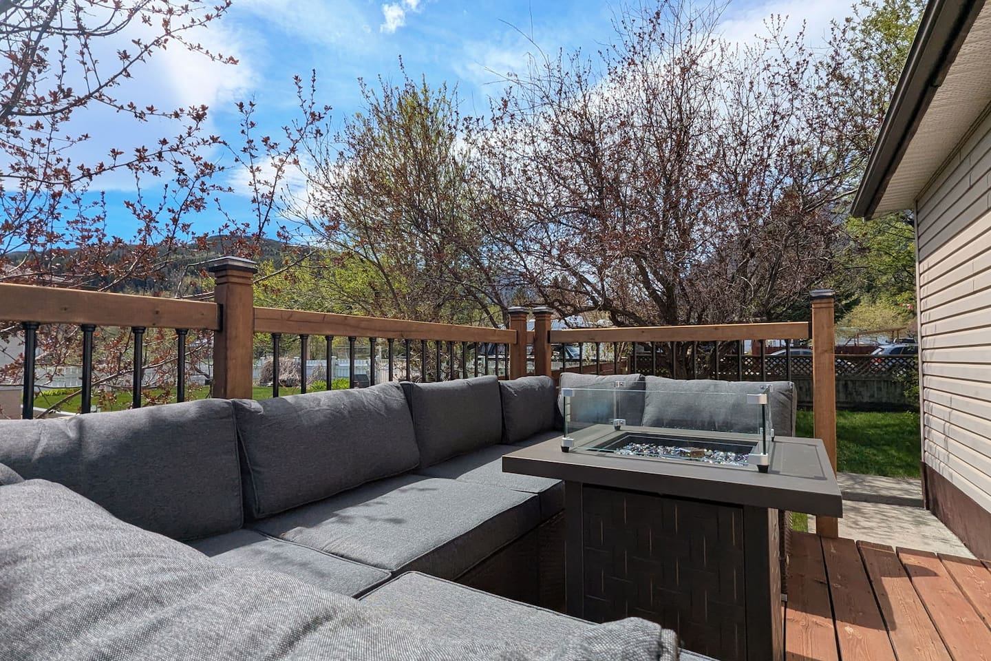 Outdoor wooden deck with a grey L-shaped sectional sofa, fire pit table, and view of distant hills.