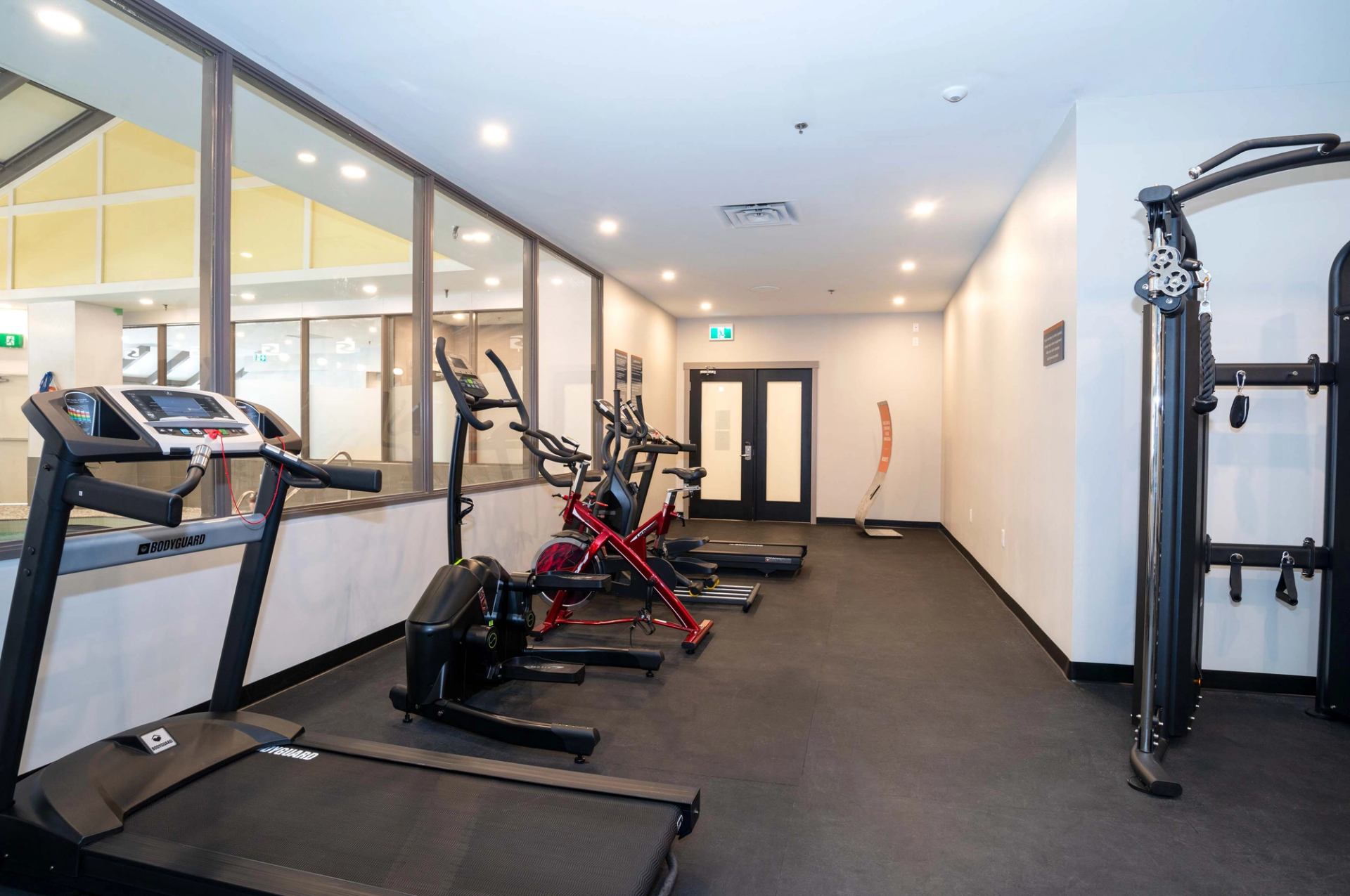 Fitness room with treadmills, stationary bikes and weight machine beside glass wall and doors.