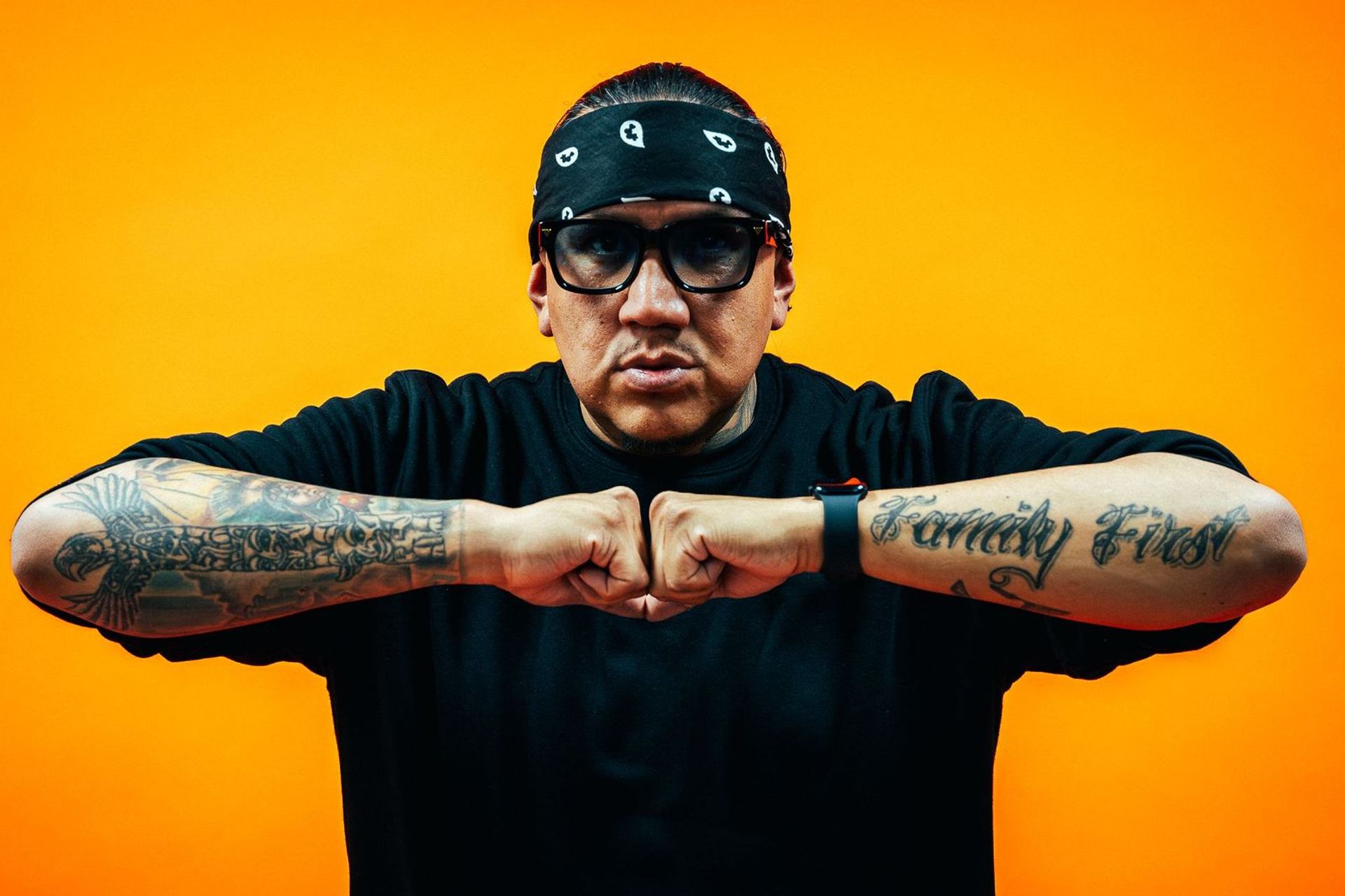 Artist with tattooed arms clasping fists against a bold orange backdrop for a live hip-hop show.