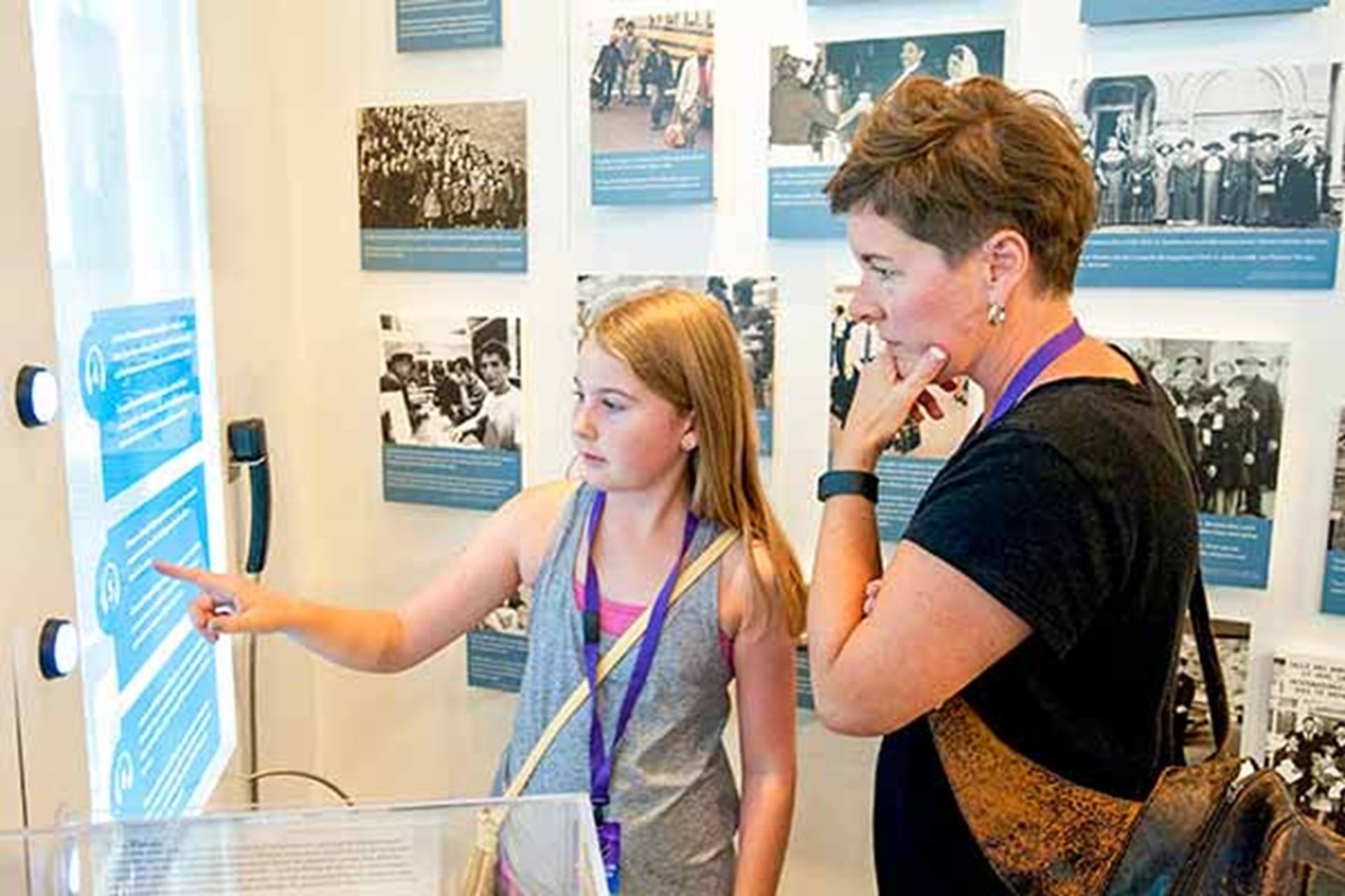 Two visitors read a touchscreen display in a museum gallery with historical photos.