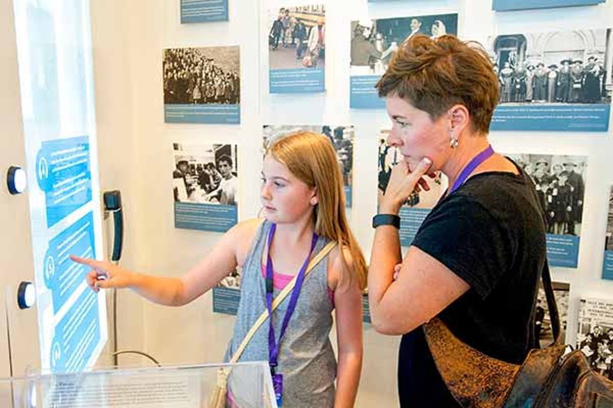 Two visitors read a touchscreen display in a museum gallery with historical photos.