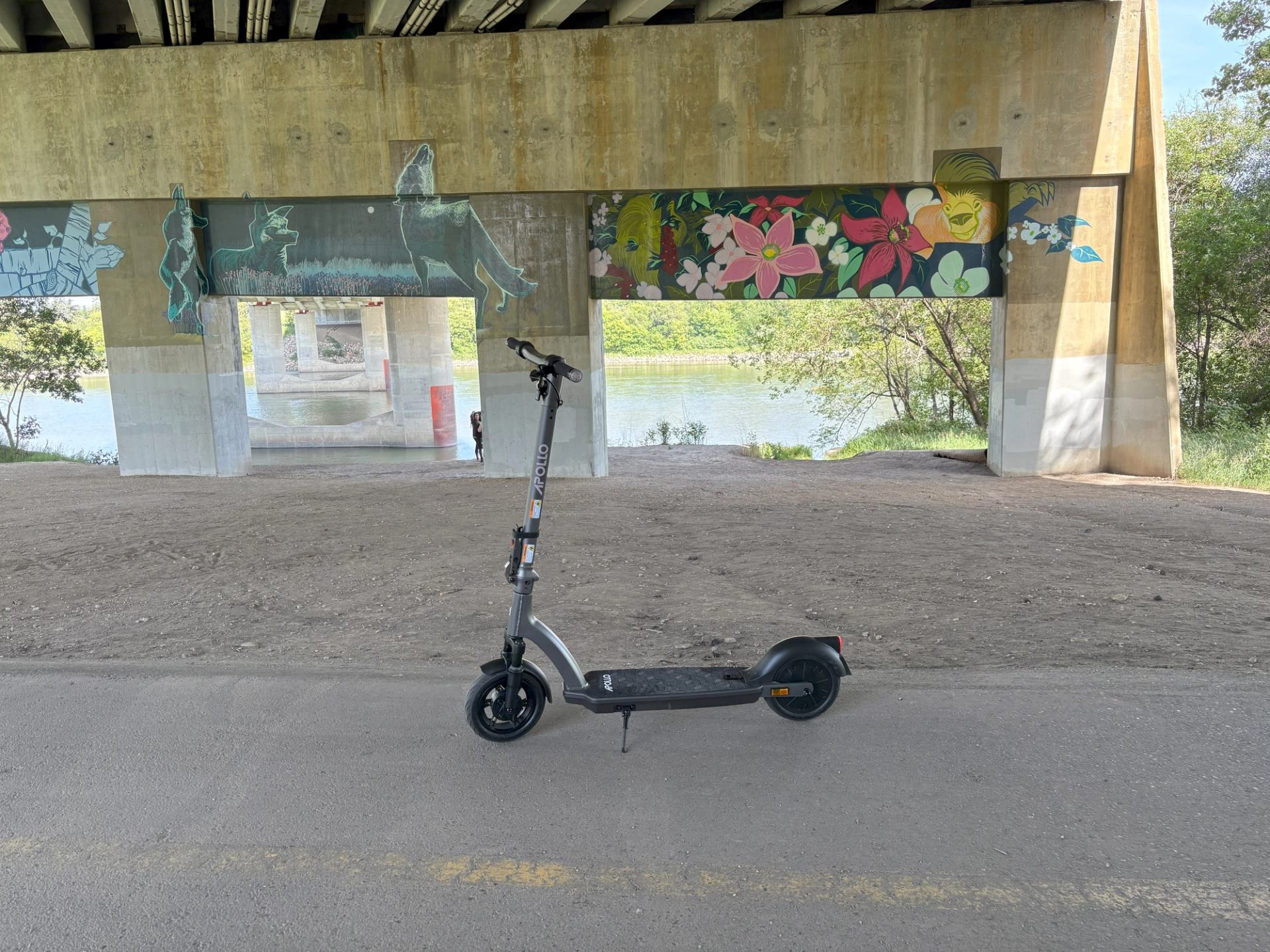 Electric scooter parked under mural-covered bridge with greenery and water behind.