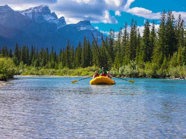 Rafters float down blue river with snowy peaks and forest all around.
