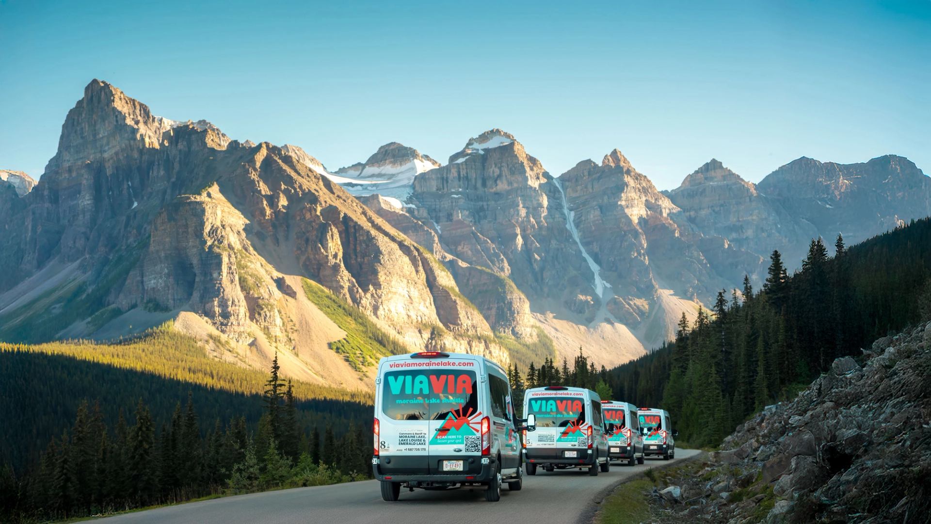 Vans on mountain road with forest, rocky terrain, and snow-capped peaks in background.