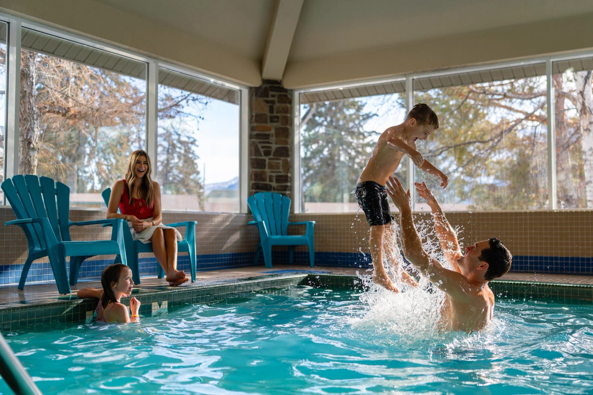 Indoor pool scene with people swimming, splashing and sitting on blue deck chairs.