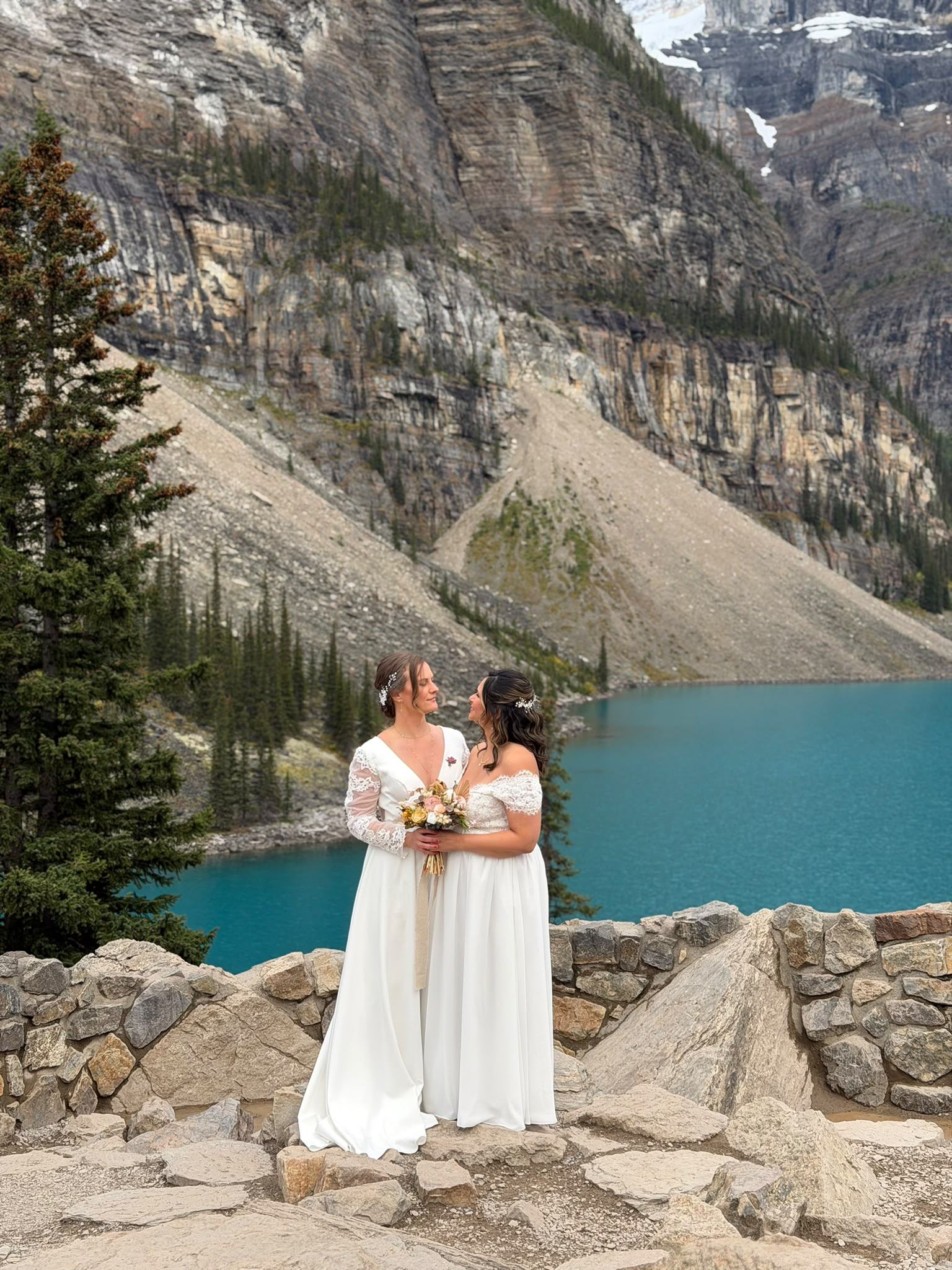 Two brides in white dresses hold a bouquet beside a turquoise mountain lake.