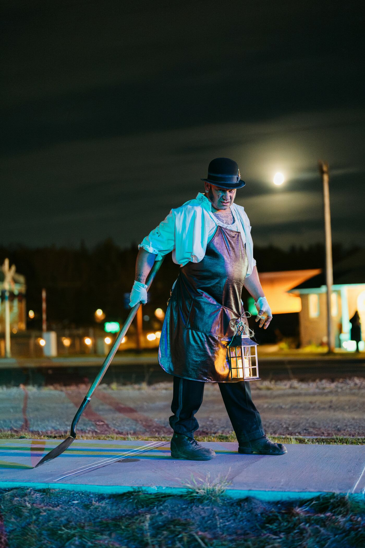 A sinister man in a bowler hat holds a lantern and scythe under a full moon