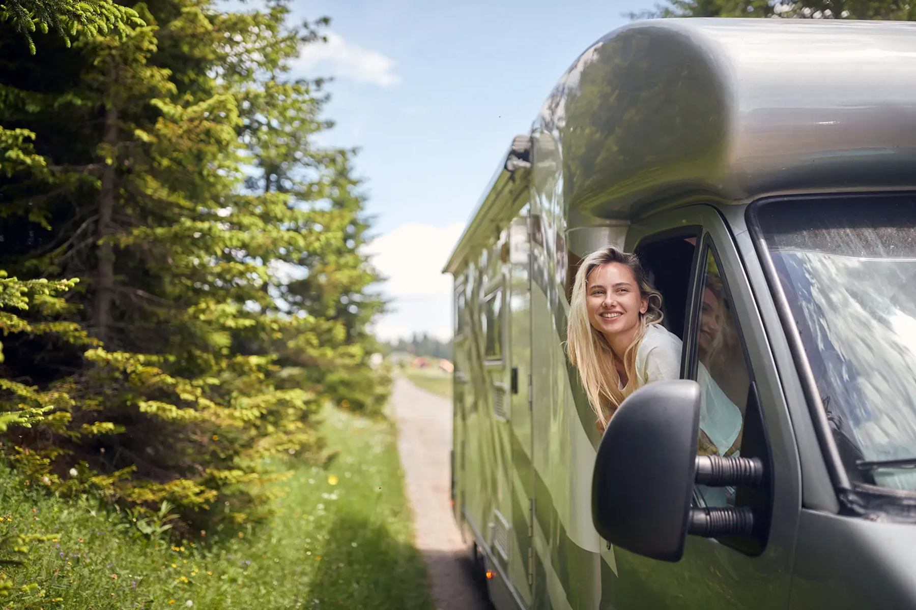 Person leaning out of camper van window on a forest road under a partly cloudy sky.