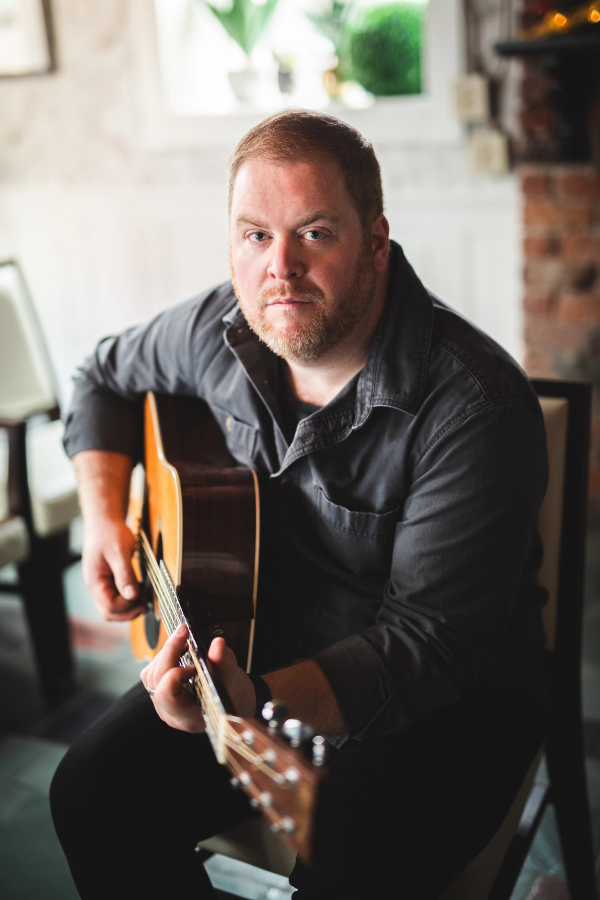 Person sitting indoors, holding an acoustic guitar and looking toward the camera.