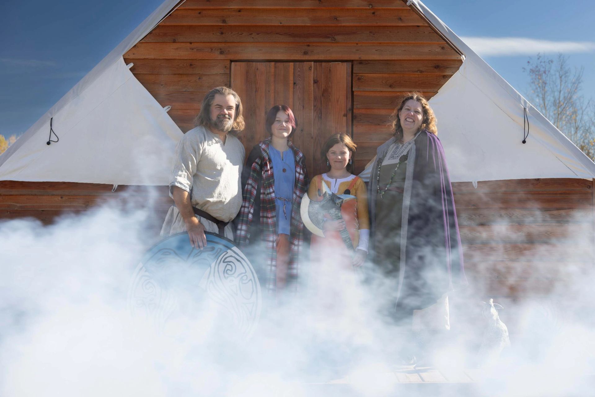 A family of four in Viking costumes, holding shields, stands in front of a rustic building, partially obscured by fog.