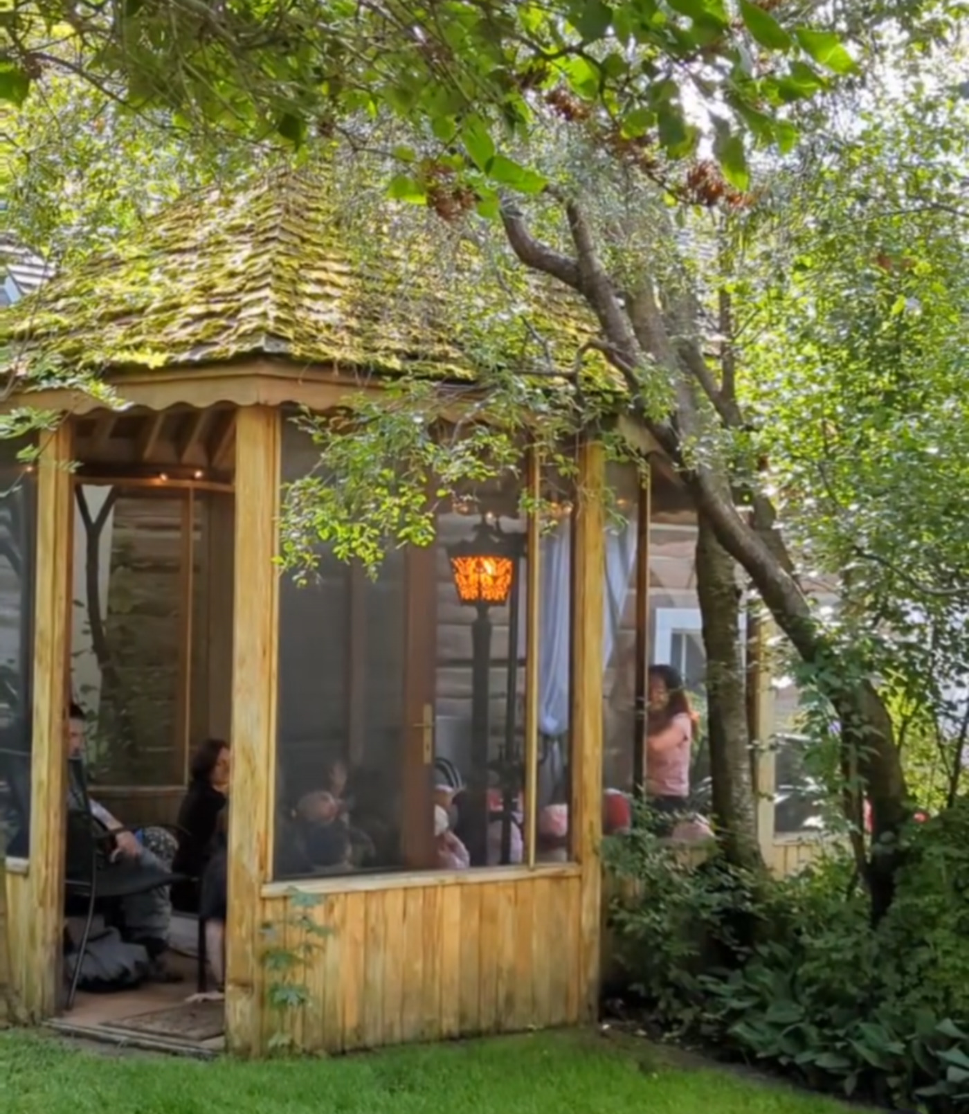 Garden walkway beside open wooden pavilion with people seated under leafy trees.