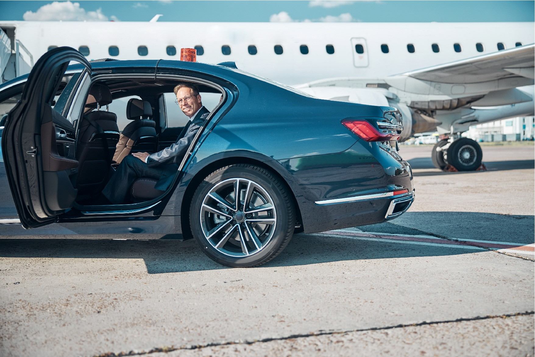 Luxury car with open rear door beside private jet on tarmac, passenger seated inside.