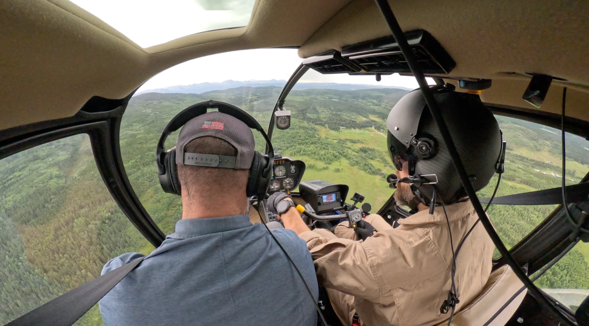 Helicopter cockpit view with two people wearing headsets flying over green forested hills.