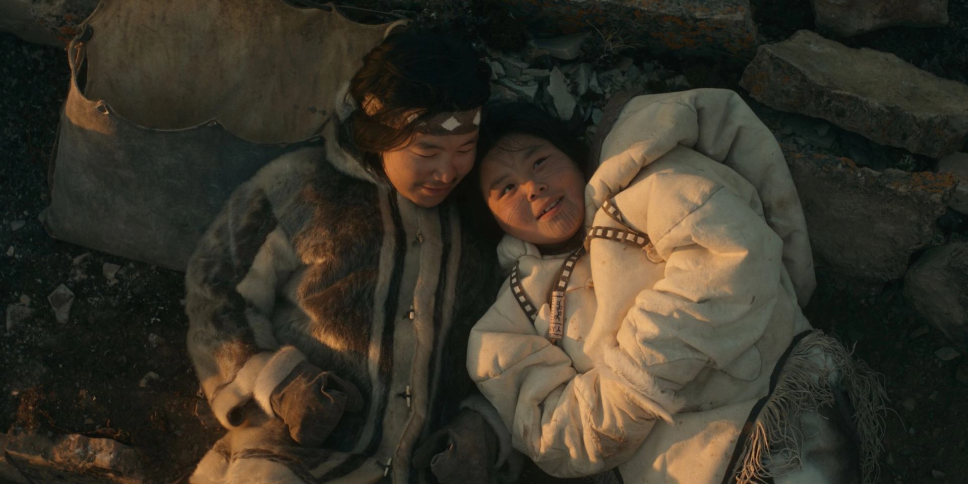 Two figures in heavy fur clothing lying close together on rocky ground in a stark Arctic landscape at dusk.