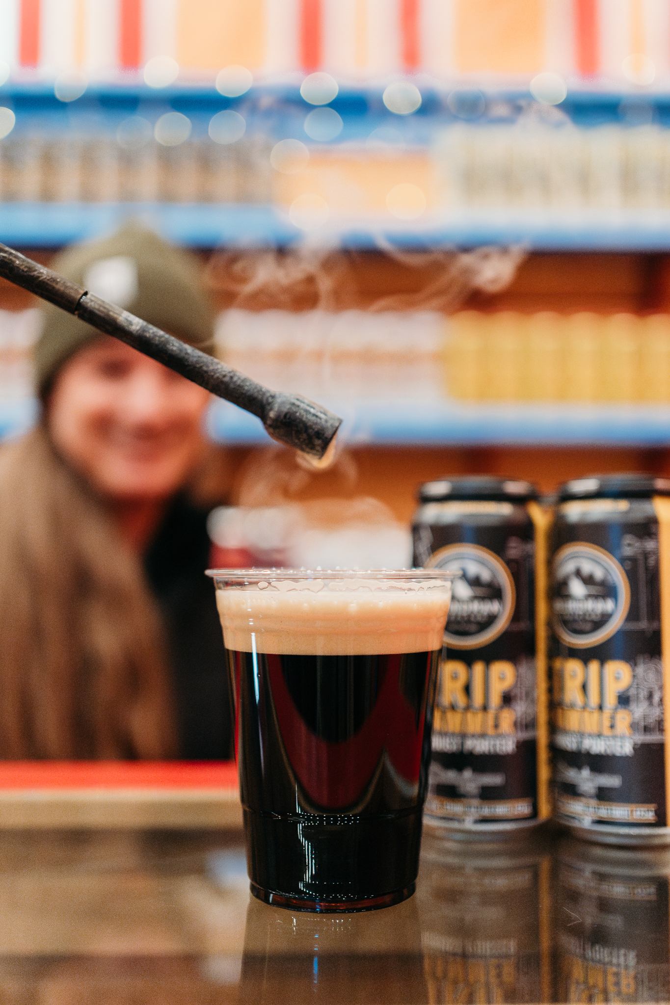 A steaming dark beverage in a clear plastic cup on a counter, with cans labeled “RIP” in the background and a torch adding heat to the drink.