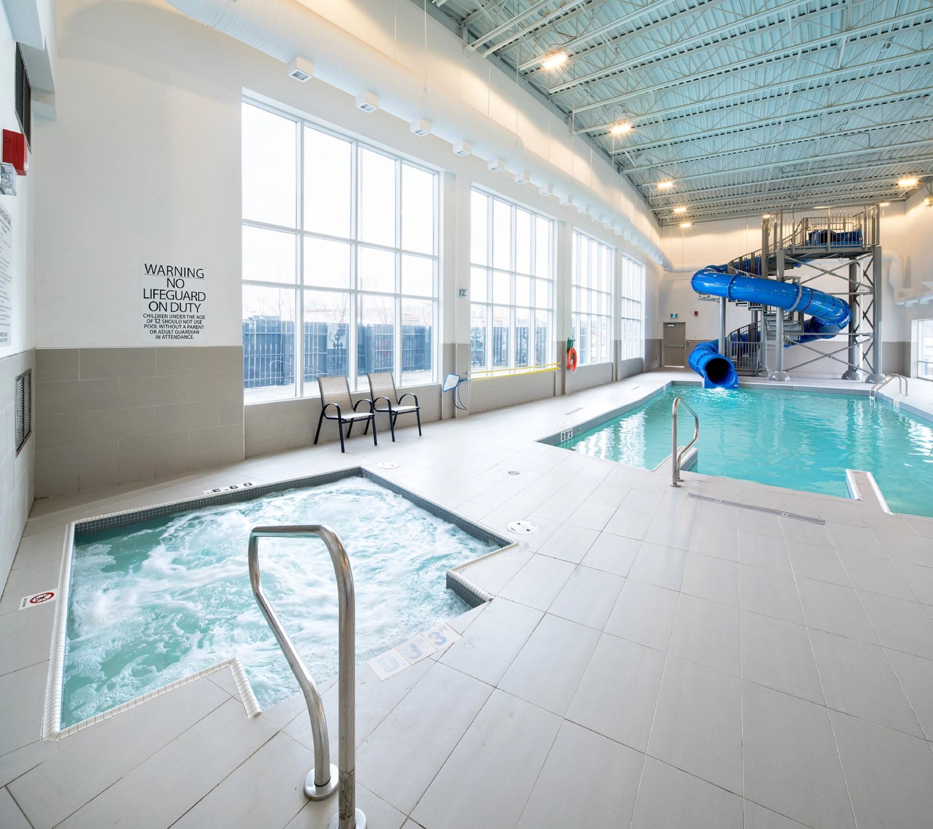Indoor pool area with a water slide, jacuzzi, and large windows; chairs are set against the wall. Bright, clean, and inviting atmosphere.