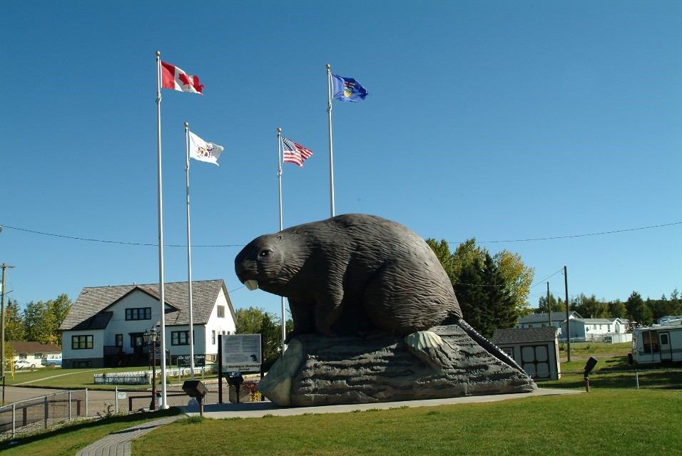 Giant beaver statue on a mound with flagpoles and small buildings behind.