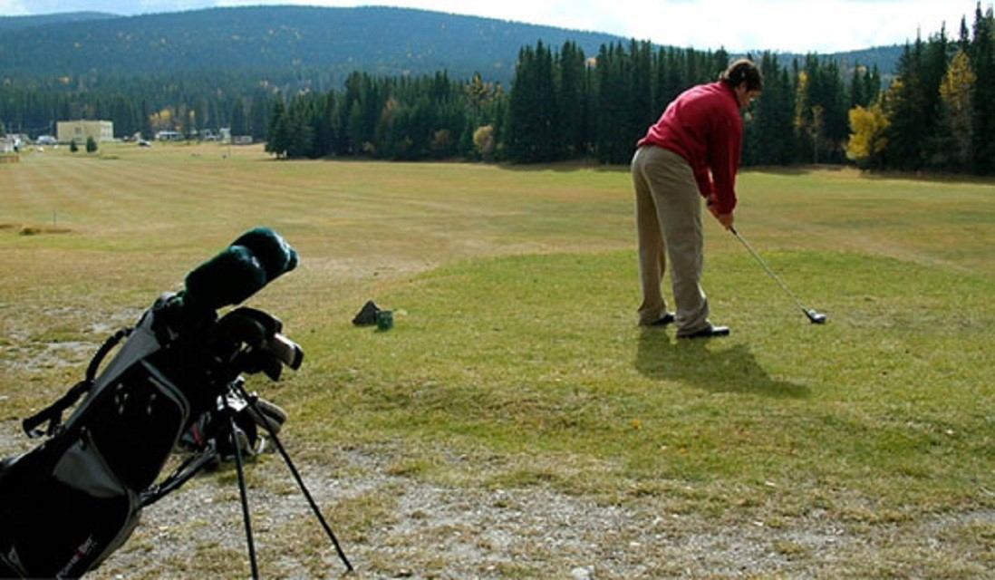 Golfer mid-swing with bag nearby, surrounded by trees and mountain views.
