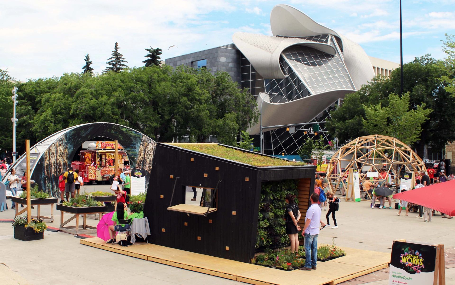 Outdoor public art installation with green-roof structure, food truck and people near modern museum.