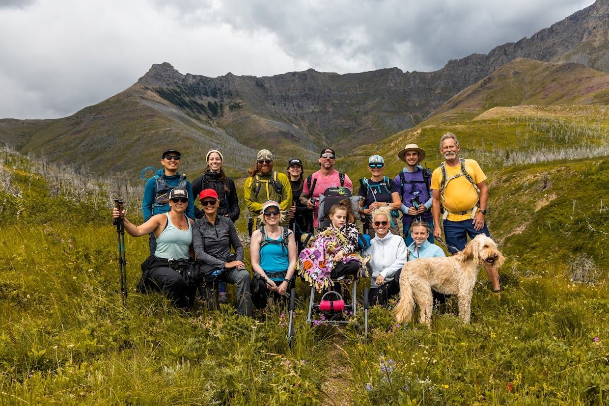 Group hiking together in a scenic mountain meadow with an adaptive participant and a dog.