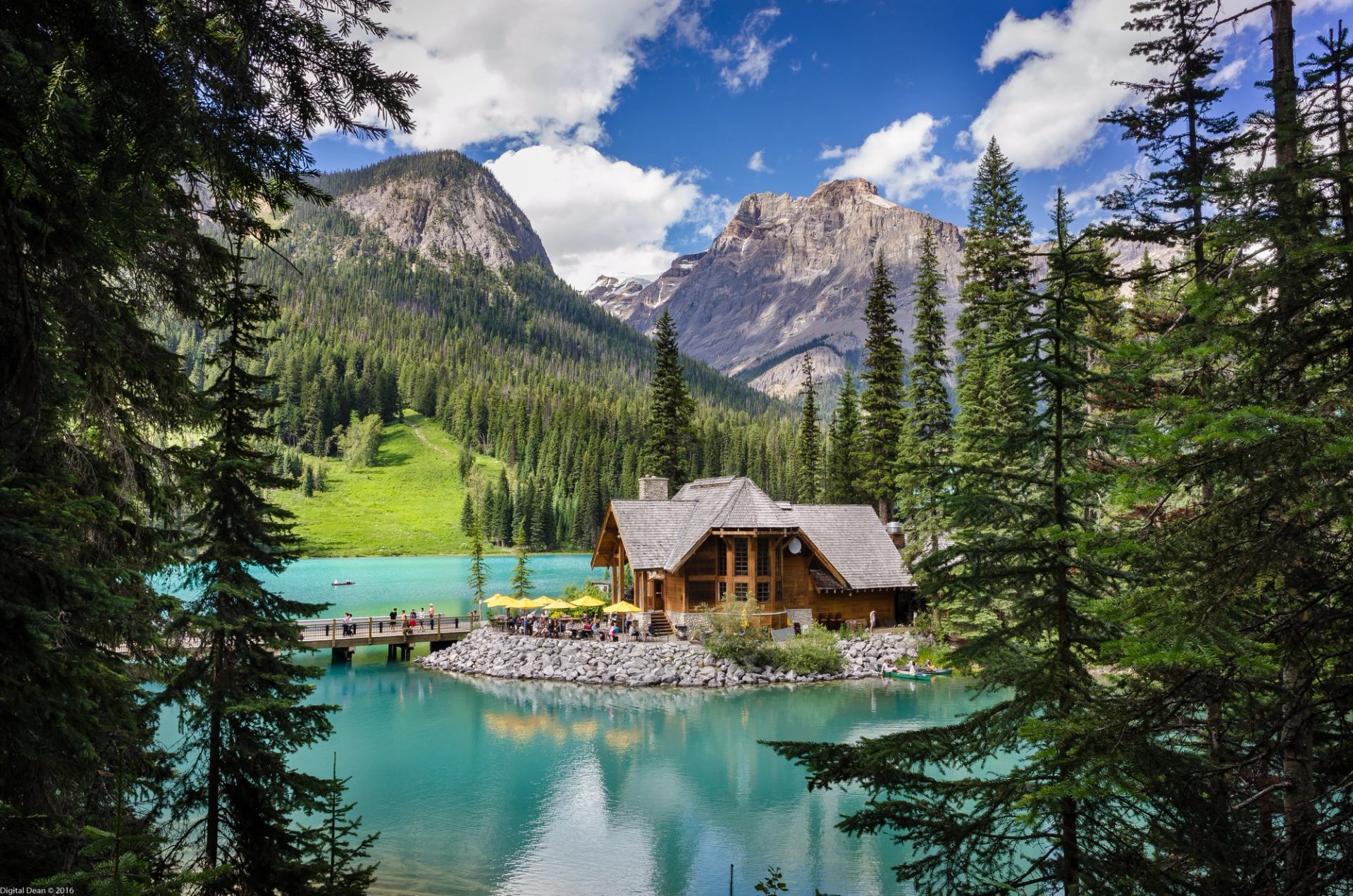 Lakeside cabin with bridge, turquoise water, forest, and mountain backdrop.