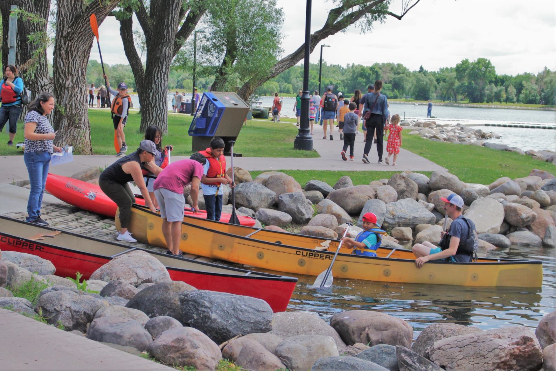 People prepare canoes at a rocky lakeshore while others walk along the park path.