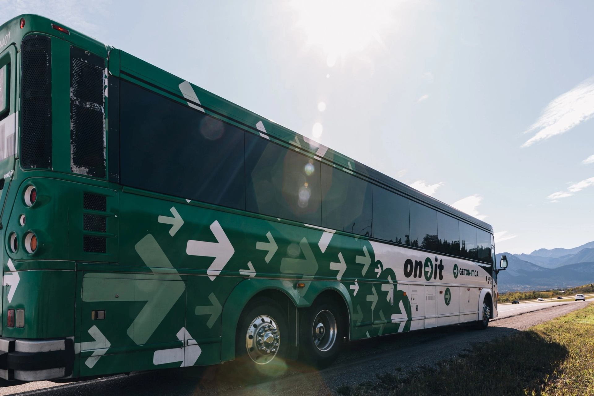 Green and white On-It bus with arrows on side, driving near mountains under a clear sky.