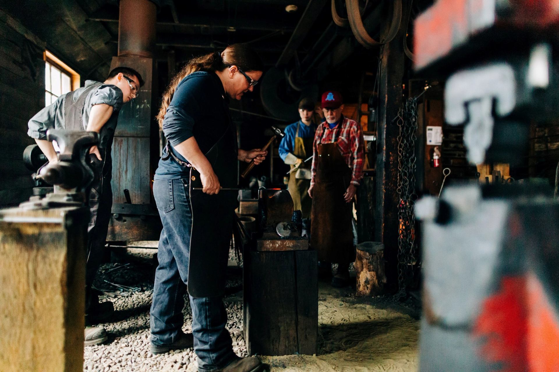 Blacksmiths work at a forge in a museum shop, with an anvil and metal tools visible.
