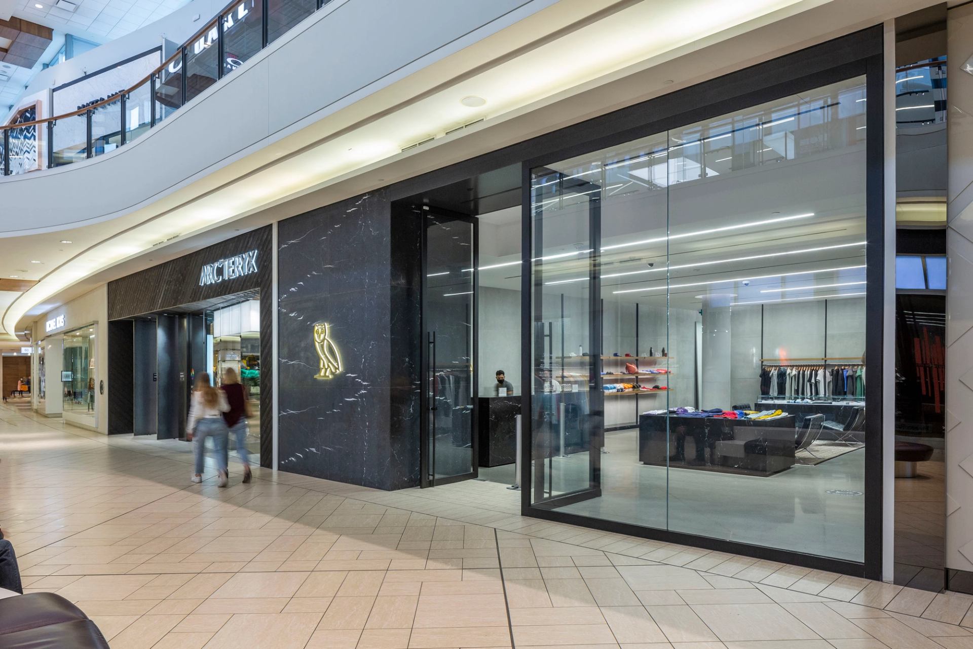 Arc'teryx storefront with glass windows in mall corridor, shoppers walking past.