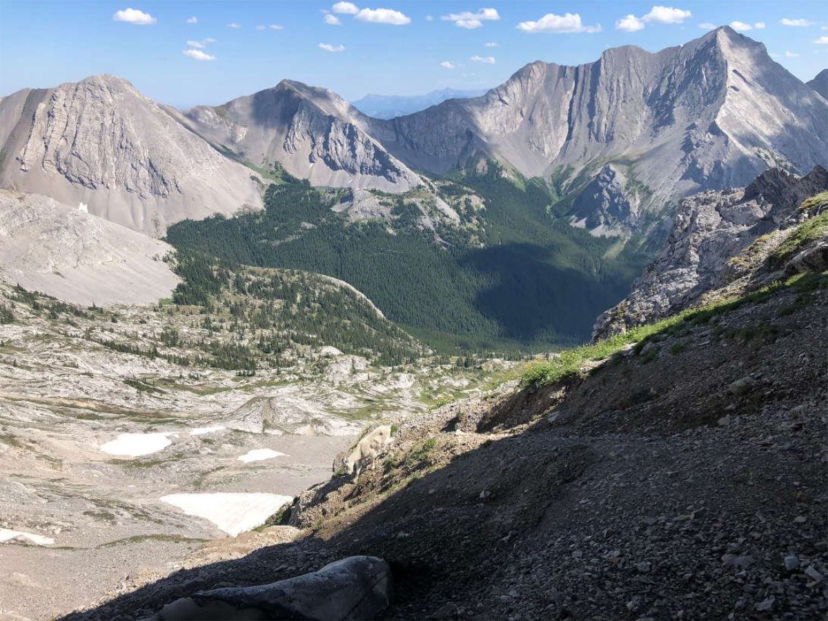 Rocky mountain valley with steep grey peaks, forested slopes and scattered snow patches.
