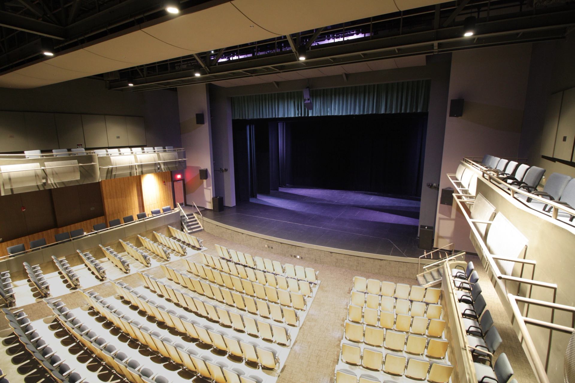 Theatre seating and stage with curtains, speakers, and overhead lighting from the balcony.