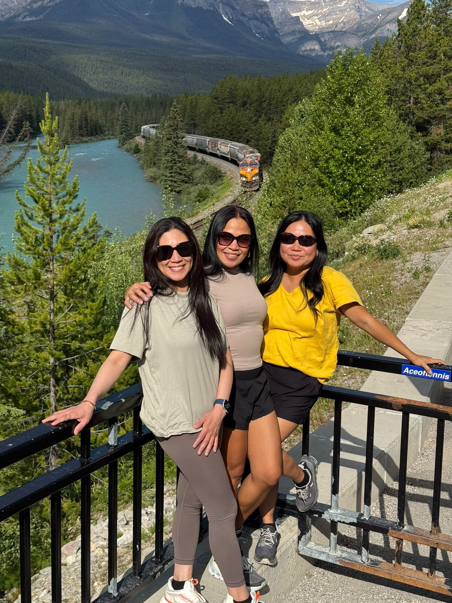 Three people pose at a lookout above a turquoise river, forest, mountains and passing train.