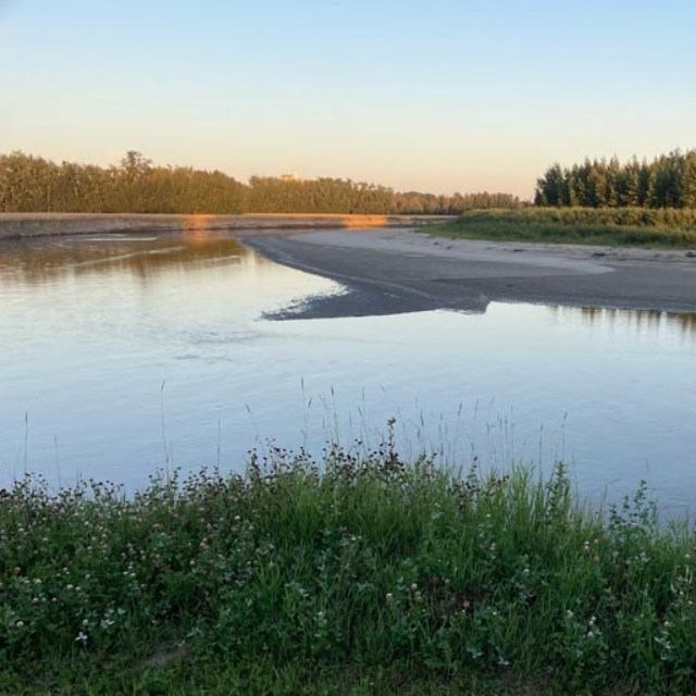 Calm river with sandy bank and green grass under a clear evening sky.