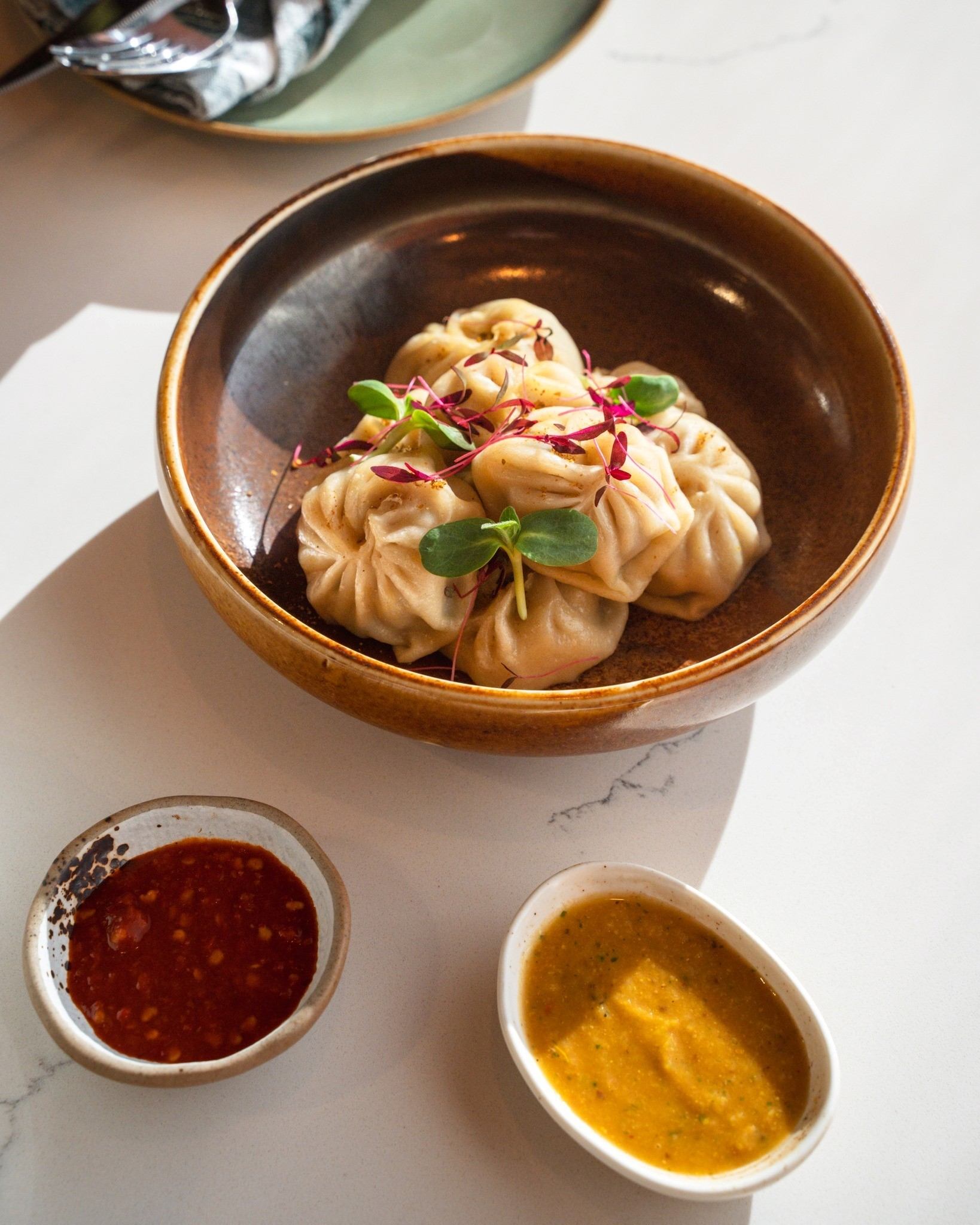 Bowl of dumplings topped with microgreens and purple sprouts, served with dipping sauces.