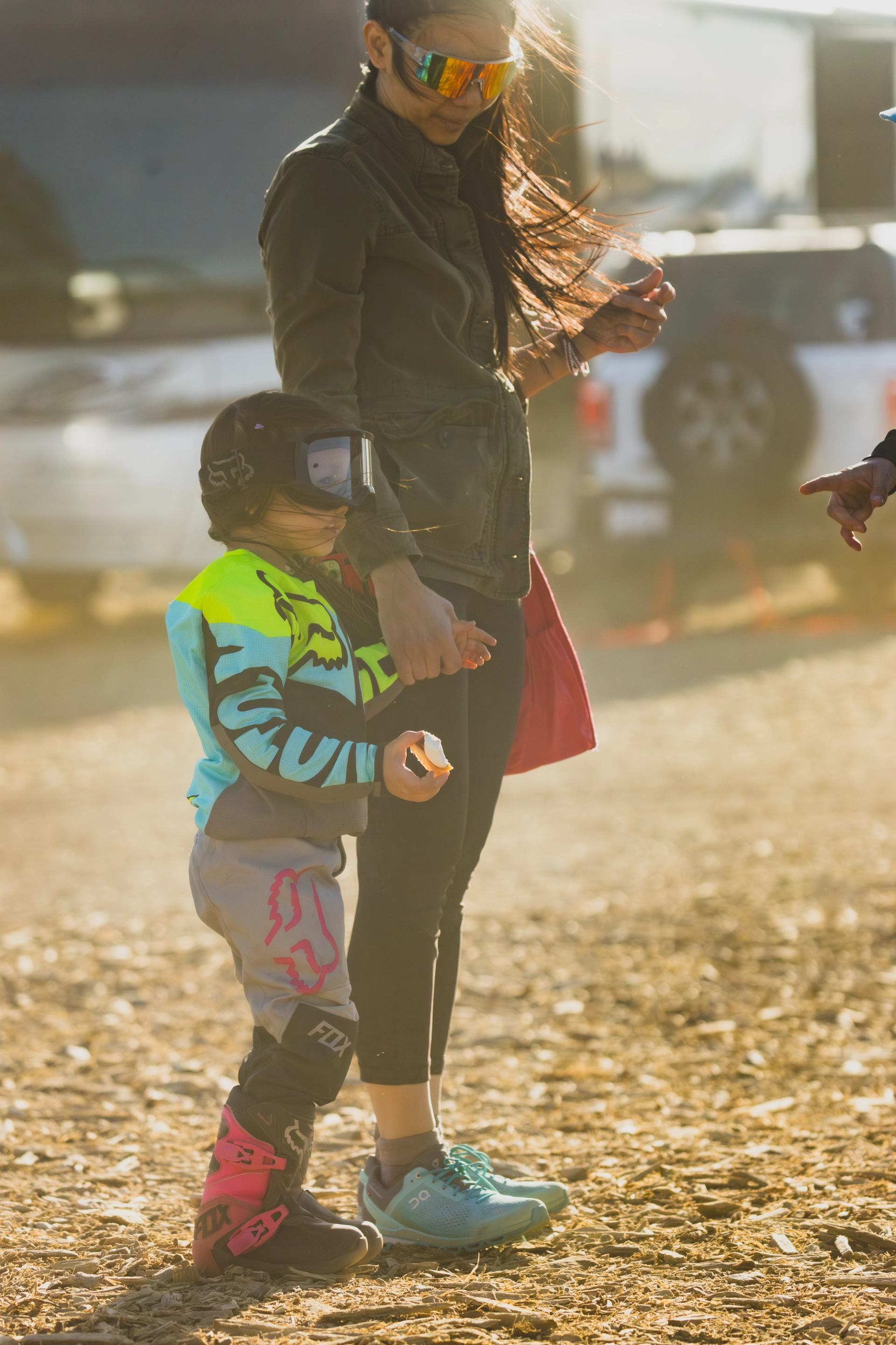 Adult and child in motocross gear standing on dirt with vehicles in background.